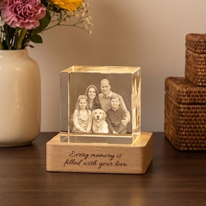 Può includere: Un cubo di vetro trasparente con un ritratto di famiglia inciso al laser insieme a un cane, illuminato da una base in legno chiaro con l'incisione "Every memory is filled with your love." Il cubo è posto accanto a un vaso di girasoli e rose.
