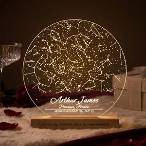 May include: A circular night light with a constellation design. The clear acrylic disc shows a star map, with the name "Arthur James" and location details. It sits on a wooden base with the inscription "You're my favorite place to come home to."