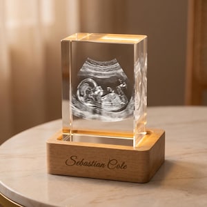 May include: A rectangular glass block featuring a laser-etched ultrasound image of a fetus inside. The glass block sits on a light-colored wooden base that is illuminated from within and displays the engraved name Sebastian Cole on the front.