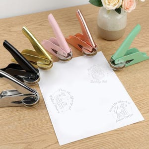 Op de afbeelding: Een verzameling kleurrijke metalen zegelpersen in zwart, zilver, goud, roze, koraal en groen. Een wit vel papier is gestempeld met diverse bibliotheekthema's en tekst. Een kleine vaas met bloemen staat op de achtergrond.