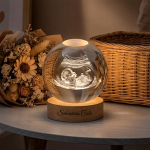 May include: A clear glass sphere with an illuminated ultrasound image of a baby, resting on a wooden base. The base is engraved with the name "Sebastian Cole". The sphere is lit from within, creating a warm glow.