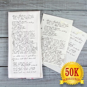 May include: A white kitchen towel and handwritten recipe cards with a recipe for "Bangue Shepherd Bread." The towel has red stripes at the bottom. A gold seal reads "50K Memories Made!"