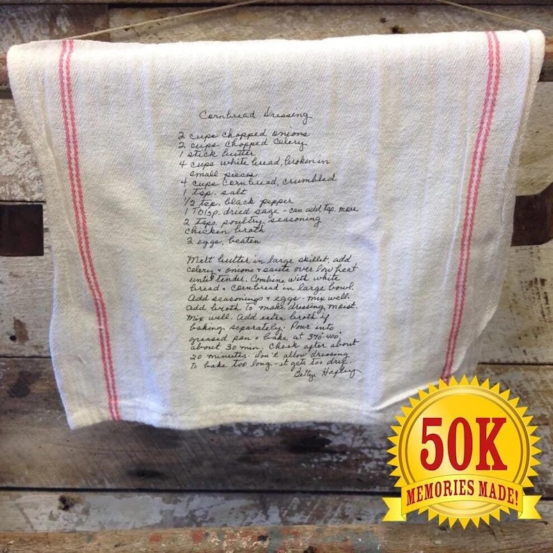 May include: A white tea towel with red stripes hangs on a wooden surface. The towel has a handwritten recipe for cornbread dressing, including ingredients like onions, celery, sage, and poultry seasoning. The recipe ends with "Bettye Haplin" and a gold star sticker with the text "50K Memories Made!"