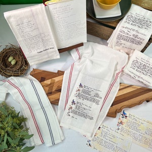 May include: Four white tea towels with red and blue stripes. Each towel features a handwritten recipe for a different dish. The recipes include "Potato Chip Cookies", "Grandma Chapman's Recipe", "Green Bean Casserole", and "Rice Krispie Treats".