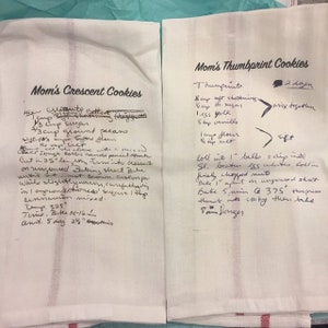 May include: Two white kitchen towels with handwritten recipes for "Mom's Crescent Cookies" and "Mom's Thumbprint Cookies." The recipes include ingredients and baking instructions. The towels have red stripes at the bottom.
