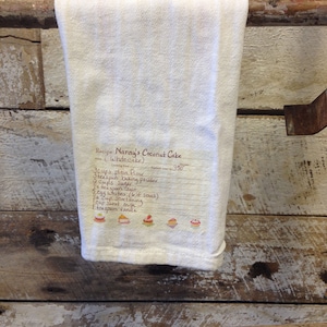 May include: A white tea towel with a handwritten recipe for Nanny's Coconut Cake. The recipe includes ingredients like plain flour, baking powder, sugar, salt, egg whites, shortening, milk, and vanilla. The towel has a decorative border of small cupcakes.