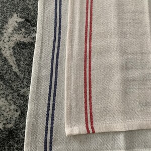 May include: Two white kitchen towels with vertical stripes. One towel has navy blue stripes, and the other has red stripes. The towels are made of a textured fabric and are folded.