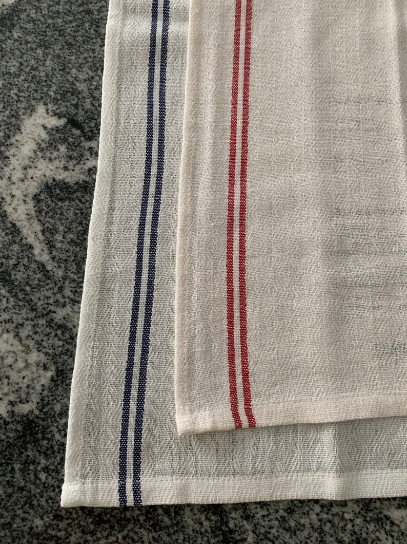 May include: Two white kitchen towels with vertical stripes. One towel has navy blue stripes, and the other has red stripes. The towels are made of a textured fabric and are folded.
