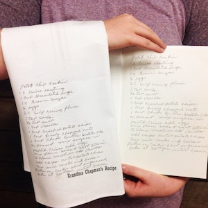 May include: A white kitchen towel and a handwritten recipe card are held up. The towel features the recipe for "Potato Chip Cookies" in black ink, along with "Grandma Chapman's Recipe" at the bottom. The recipe card has the same recipe.
