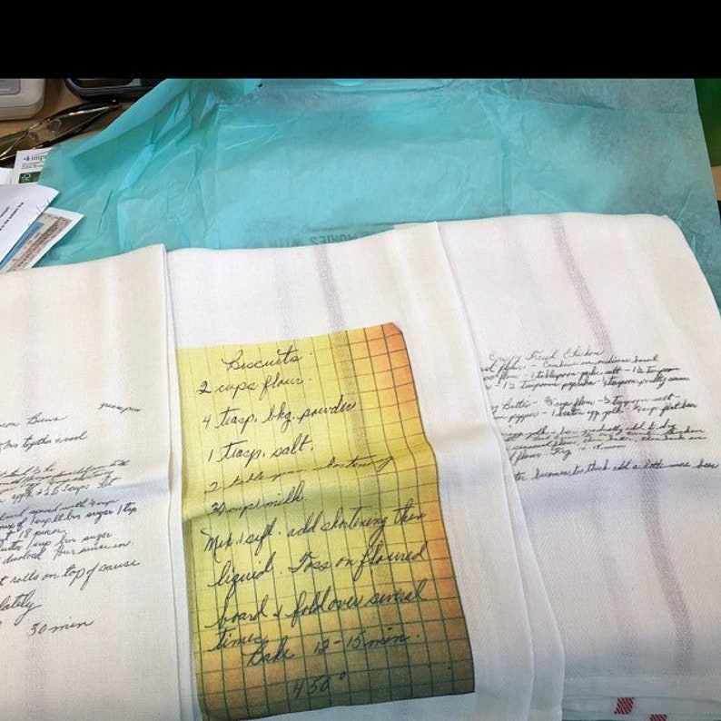 May include: White kitchen towels with red stripes, featuring handwritten recipes. One towel displays a recipe for biscuits, including ingredients like flour and baking powder. The towels are folded and arranged on a light blue surface.