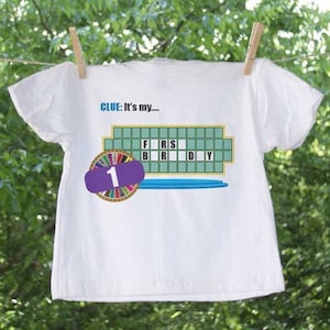 May include: White t-shirt with a Wheel of Fortune game board design. The board shows the letters "F, R, S, B, R, D, Y" and the clue "It's my..." is printed above the board. A purple wheel with the number "1" is on the left side of the board.