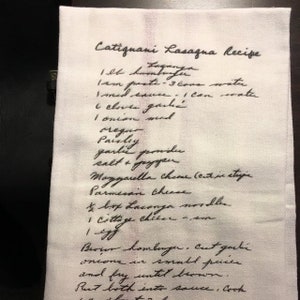 May include: A white kitchen towel with a handwritten lasagna recipe in black ink. The recipe includes ingredients like lasagna noodles, ground beef, and mozzarella cheese. The towel is folded and has a slightly textured surface.