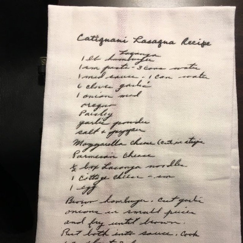 May include: A white kitchen towel with a handwritten lasagna recipe in black ink. The recipe includes ingredients like lasagna noodles, ground beef, and mozzarella cheese. The towel is folded and has a slightly textured surface.