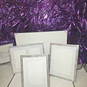 May include: Four white picture frames with silver rhinestone borders are displayed on a white surface. The frames have white matting and are empty. A sign with the text "Bling Therapy" is visible in the background. The background is a shiny purple material.
