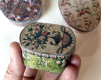 May include: Three small decorative metal boxes with lids. One is round with a brown floral pattern, one is oval with a light floral design, and one is rectangular with rounded corners, featuring a green base and a floral urn graphic on top.
