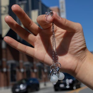 May include: A silver necklace with multiple charms and white earbud-style attachments is held up against a bright blue sky. The necklace is held by a hand, with a blurred background of buildings and cars.