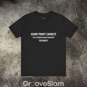 May include: Black t-shirt with white text: "SENIOR PROMPT ENGINEER I TALK TO ROBOTS BECAUSE HUMANS ARE TOO BUGGY". The short-sleeved tee has a crew neck. The brand name "GrooveSlam" is printed at the bottom of the shirt.