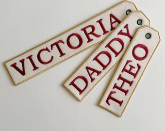 May include: Three rectangular off-white fabric tags with gold-colored stitched borders and dark red embroidered names. The tags feature the names VICTORIA, DADDY, and THEO in capital letters, each with a small metal eyelet at the top for attachment.