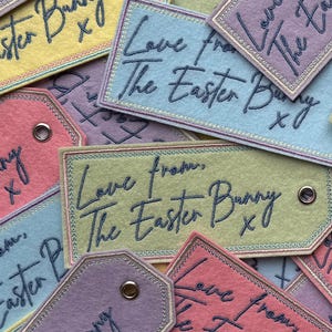 Może przedstawiać: Kolekcja kolorowych filcowych zawieszek na prezenty w pastelowych odcieniach. Każda zawieszka jest haftowana napisem "Love from The Easter Bunny xoxo" w ozdobnej czcionce, idealna na prezenty wielkanocne.