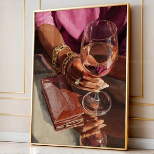 Op de afbeelding: Een ingelijste kunstdruk toont een persoon die een wijnglas met een lichtgekleurde vloeistof vasthoudt. Een bruine leren portemonnee en een gouden armband zijn ook zichtbaar. Het kunstwerk heeft een gouden lijst en een roze achtergrond.