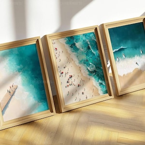 Może przedstawiać: Trzy oprawione wydruki przedstawiające sceny z plaży. Wydruki przedstawiają widoki z lotu ptaka na plaże z turkusową wodą, piaszczystymi brzegami i ludźmi. Ramki wykonane są z jasnego drewna. Wydruki artystyczne oparte są o białą ścianę na drewnianej podłodze.