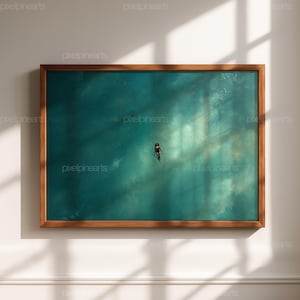 May include: A framed art print featuring a person swimming in turquoise water. The artwork is set in a wooden frame and is mounted on a white wall. The image is lit by sunlight, creating shadows.