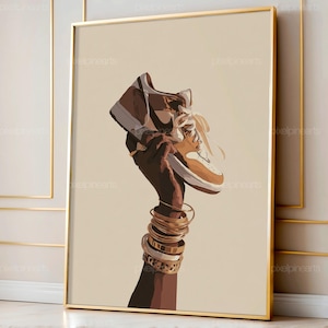 May include: A framed art print depicting a brown and tan sneaker held aloft by a dark-skinned hand, embellished with gold bangles. The sneaker has white laces and a brown swoosh. The background is a plain beige.
