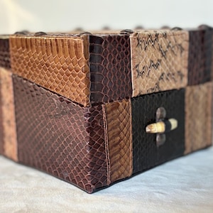 May include: A small, square box made of brown, tan, and black leather with a bone-like handle. The box is made of different pieces of leather sewn together.
