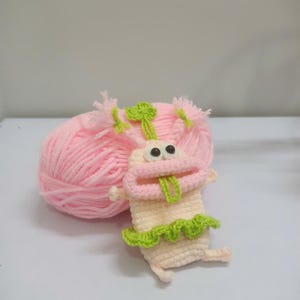 May include: A handmade crochet creature with a light pink body, a pink mouth, and a green tongue. It has a green frill around the bottom and pink yarn hair. A ball of pink yarn sits behind the creature.