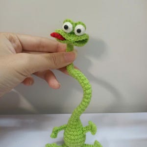 Puede incluir: Una rana de ganchillo verde hecha a mano con grandes ojos blancos y negros y una lengua roja. La rana tiene un cuello largo y curvado y brazos y piernas pequeños. Es un artículo artesanal caprichoso.
