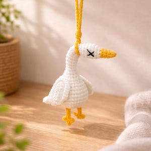 Könnte beinhalten: Eine handgefertigte weiße Häkelgans mit gelbem Schnabel und Füßen sowie einem schwarzen 'X' als Auge. Die Gans wird an einer gelben Kordel aufgehängt. Das Bastelobjekt wird vor einem weichen, verschwommenen Hintergrund präsentiert.