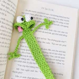 Può includere: Un segnalibro a forma di rana realizzato all'uncinetto in verde brillante, con grandi occhi bianchi, pupille nere e una piccola lingua rosa, appoggiato su un libro aperto con testo in vietnamita. Il segnalibro ha un corpo lungo e sottile.