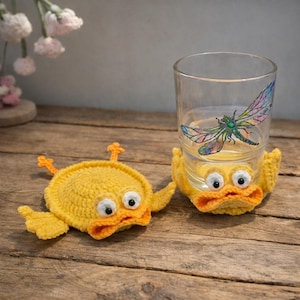 Peut inclure: Deux dessous de verre en crochet jaune en forme de canard avec des becs orange et des yeux blancs. Un dessous de verre est à côté d'un verre transparent avec un motif de libellule. Les dessous de verre sont sur une surface en bois.