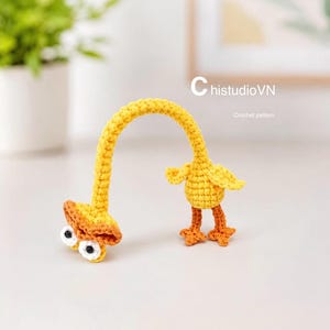 Puede incluir: Una figura de pato tejida a ganchillo en color amarillo con un cuello largo y curvado, patas naranjas y dos ojos grandes blancos con pupilas negras. El texto ChistudioVN Crochet pattern aparece en la esquina superior derecha con una C.