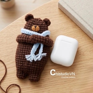 May include: A dark brown crocheted bear figure wearing a light blue scarf, positioned next to a white wireless earbud charging case. The text ChistudioVN Crochet pattern is printed in white near the bottom right of the bear figure.