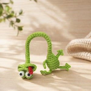 Könnte beinhalten: Zwei handgefertigte grüne Häkelfrosch-Spielzeuge. Ein Frosch hat einen langen, gebogenen Hals mit einem Gesicht und Kulleraugen. Der andere Frosch befindet sich in einer Sprungpose. Das Maul des Frosches ist mit einer roten Zunge geöffnet.