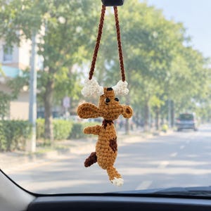May include: A brown crocheted dog ornament hanging from a rearview mirror by a brown yarn cord. The dog has a dark brown patch on its side, a white-tipped tail, and is holding a white crocheted bone above its head with its front paws.