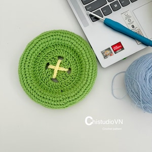 May include: A round, lime green crocheted button with a yellow cross-stitched center, positioned next to a blue crochet hook, a laptop keyboard, and a ball of light blue yarn. The text ChistudioVN Crochet pattern is visible at the bottom.