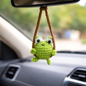May include: A handmade, green crochet frog car charm hanging from a brown cord. The frog has large, white-rimmed eyes and is suspended in front of a car's interior, adding a whimsical touch to the vehicle's decor.