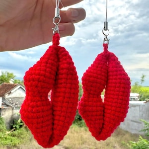 May include: Two handmade, bright red crochet lip-shaped charms. Each charm is suspended by a silver-toned clasp and a thin white cord. The background shows a cloudy sky and green foliage.