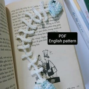 Op de afbeelding: Een gehaakte visgraat bladwijzer in wit en lichtblauw, liggend op een open boek. De bladwijzer heeft een gedetailleerd visgraatontwerp met een zachte, getextureerde uitstraling. De tekst "PDF English pattern" is zichtbaar.