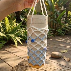 May include: A beige cylindrical container with a white and light blue crocheted holder, adorned with small blue and yellow pom-poms. The holder has a woven net design and a white rope handle. The container is held up by a hand.