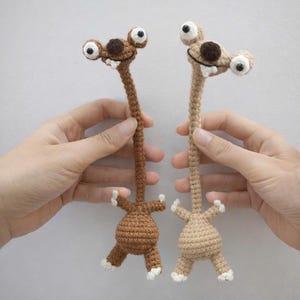 Könnte beinhalten: Zwei handgefertigte Häkeltiere, eines braun und eines beige, werden gezeigt. Jedes hat einen langen Hals, große Kulleraugen und einen kleinen Körper mit winzigen Armen und Beinen. Die Kreaturen haben weiße Akzente an Füßen und Zähnen.