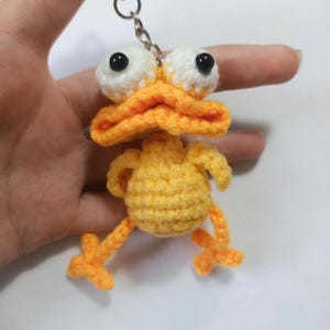 Könnte beinhalten: Ein handgefertigter gelber Häkelenten-Schlüsselanhänger mit orangefarbenem Schnabel, Füßen und Beinen. Die Ente hat große weiße Augen mit schwarzen Pupillen. Der Schlüsselanhänger ist an einem silbernen Metallring befestigt.