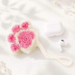 May include: A cream and pink crocheted cat paw case with a matching wrist strap, positioned next to a white wireless earbud charging case and a single white earbud. The text ChistudioVN is visible in the lower center of the image.