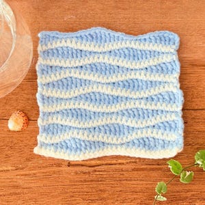 Puede incluir: Un posavasos cuadrado, tejido a mano con ganchillo, con un patrón ondulado en hilo azul claro y blanco. El posavasos está sobre una superficie de madera, con un cuenco de cristal y una pequeña concha cerca. El diseño del posavasos se asemeja a las olas.