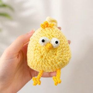 May include: A small, round, yellow crocheted chick toy featuring a textured yarn body, two large white eyes with black pupils, a small orange beak, a tuft of yellow feathers on top, and two thin yellow legs with three-toed feet dangling below.