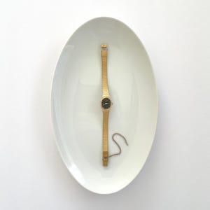May include: A gold-toned wristwatch with a black watch face rests on a white, oval-shaped dish. The watch has a delicate, woven band. A small, detached chain lies near the watch. The watch and dish are set against a white background.