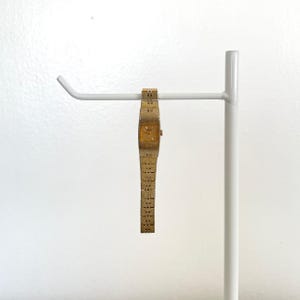 May include: A gold-toned vintage wristwatch with a rectangular face and a textured band. The watch is suspended on a white metal display stand. The watch face has a gold color and a small dial.