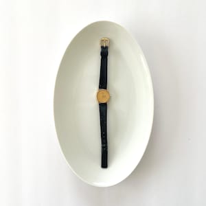 May include: A gold-toned wristwatch with a dark blue band and buckle rests on a white, oval-shaped dish. The watch face is circular, and the band is thin. The dish is empty and has a smooth surface.